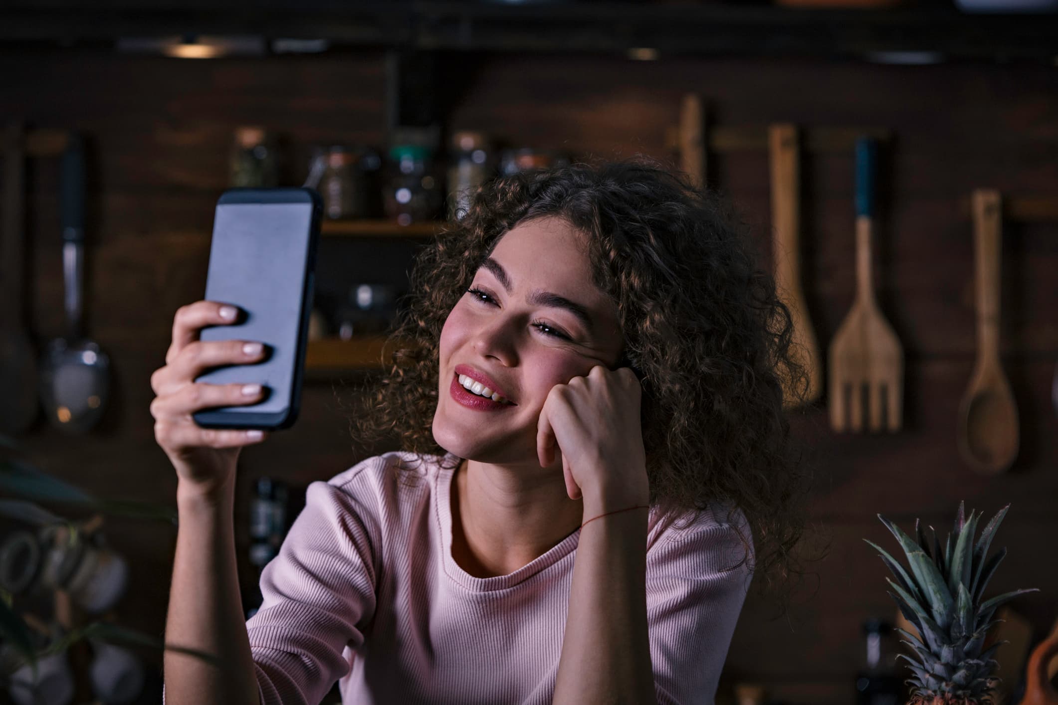 A young woman, with curly hair parted in the centre and reaching down to her chin, smiles as she holds a smartphone in her right hand