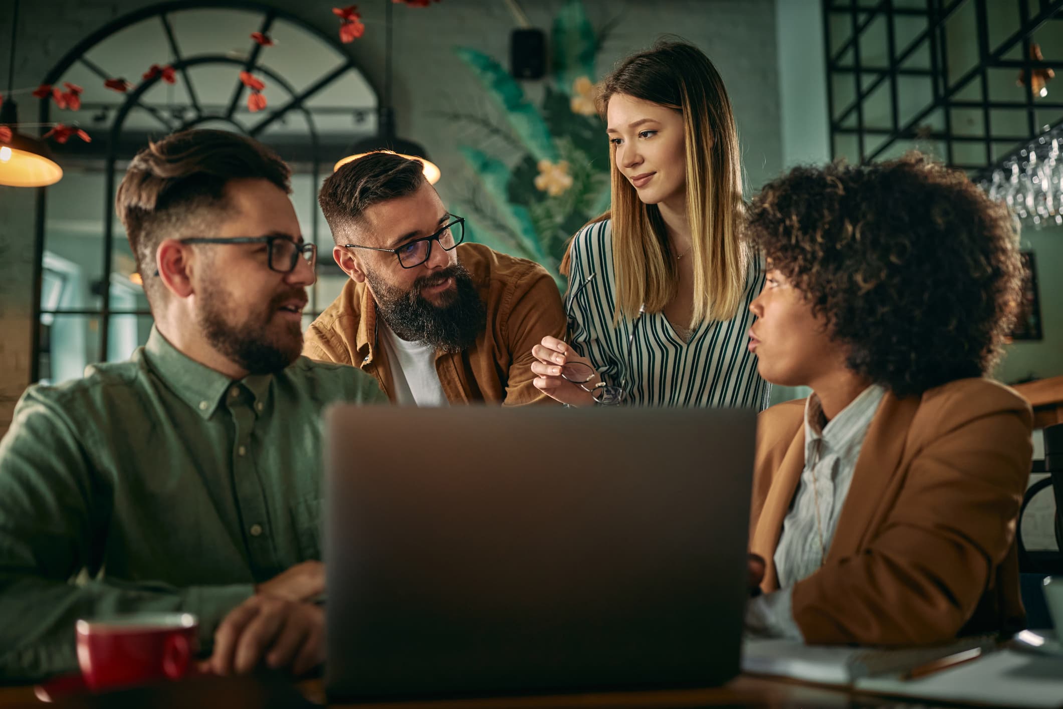 Two beared and bespectacled men behind a laptop face a woman to their left while a woman to their right looks toward them.