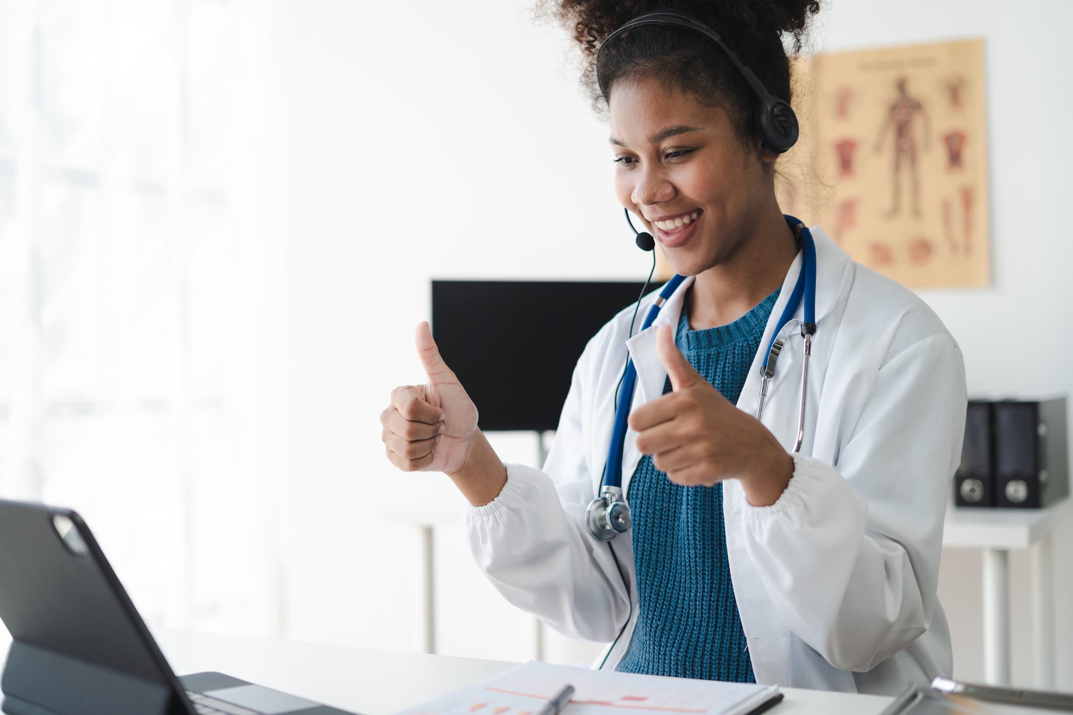A female doctor wearing headphones puts both of her thumbs up as she stands and looks at a laptop.