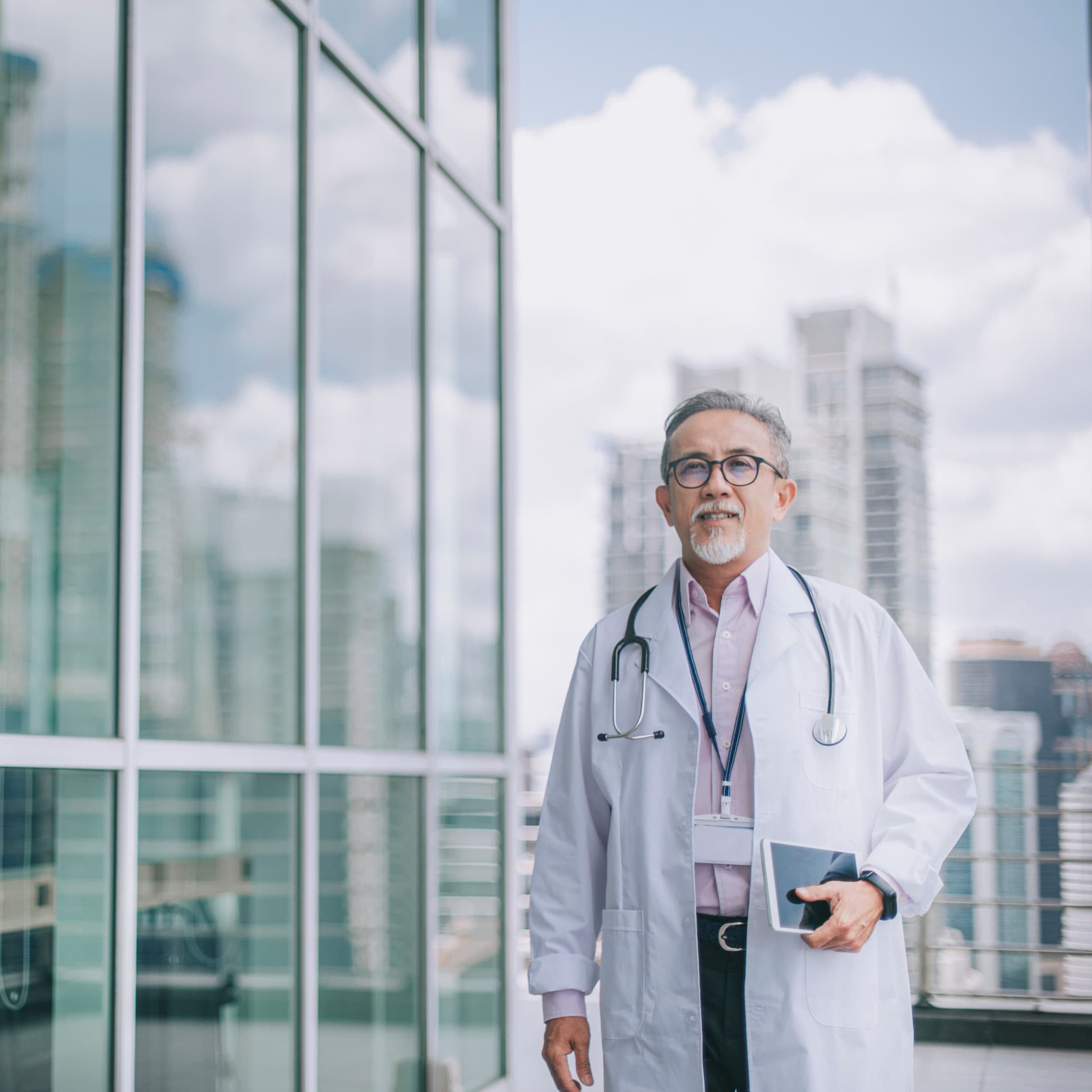 A bespectacled doctor with a goatee beard is outside with skyscrapers and a cloudy sky behind him.
