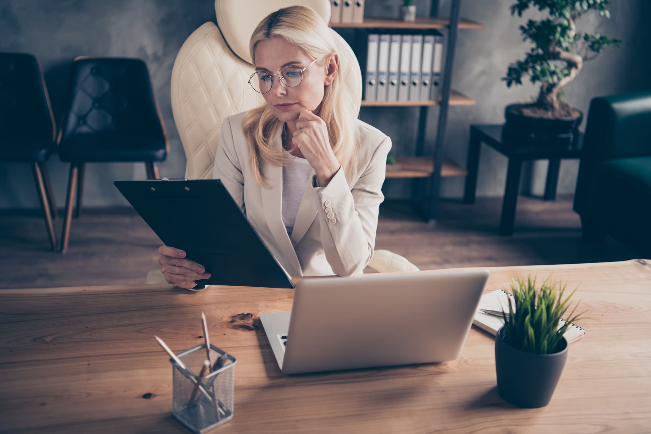 A woman wearning glasses and a smart blazer sits in a chair, in front of a laptop, a pot of pens, a plant and a notepad, looking at a clipboard held in her right hand.
