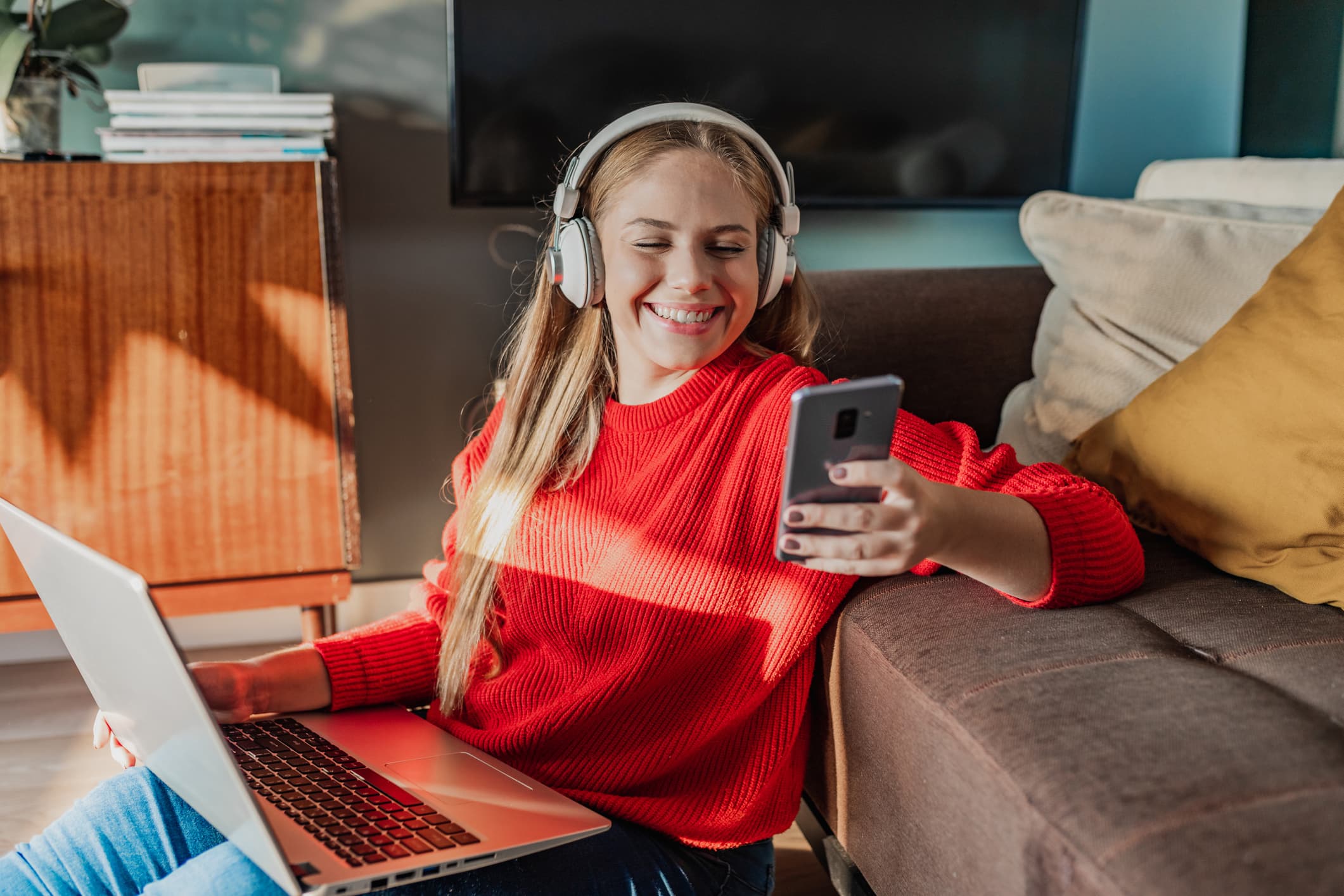 A woman wearing headphones holds a smartphone in her left hand and a laptop in her right hand, as she sits on the floor, with her back resting against a couch.