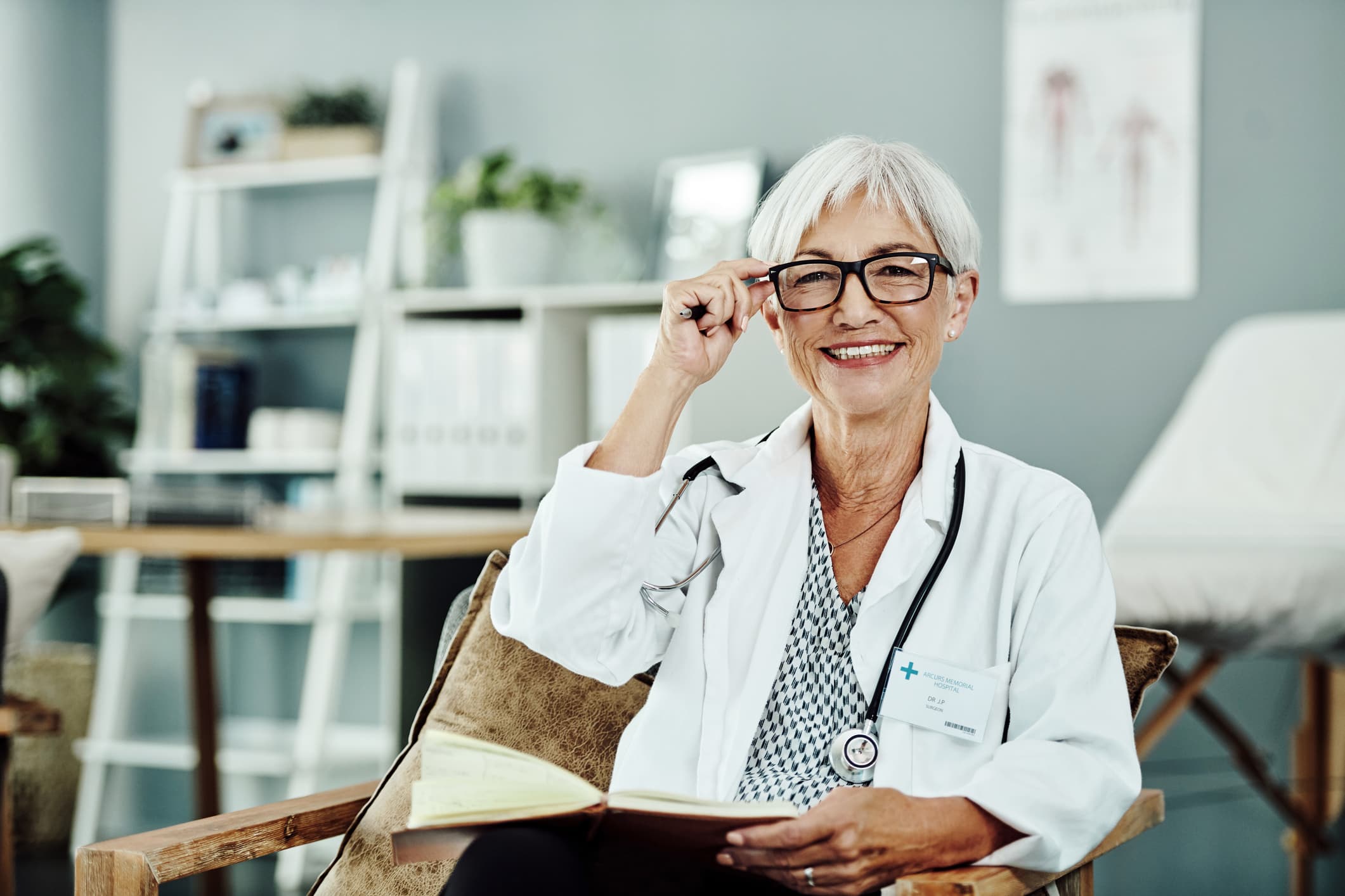 A woman wearing a doctors coat looks at you while adjusting her glasses with her right hand and holding a notebook with her left hand.
