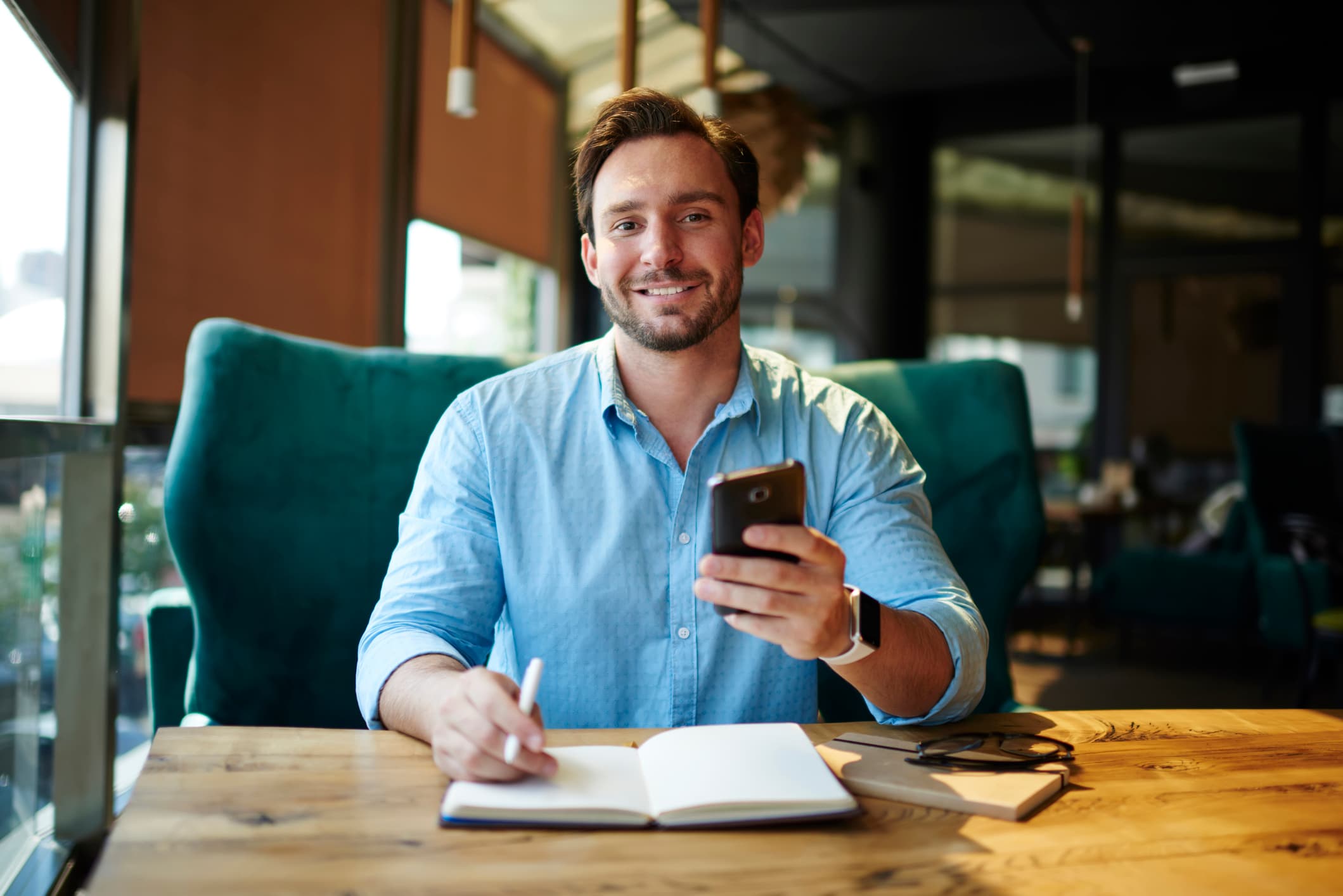 A man sits in a chair, at a desk, facing the camera while he holds a phone in his left hand and a pen in his right hand. His right hand is resting on a notebook.