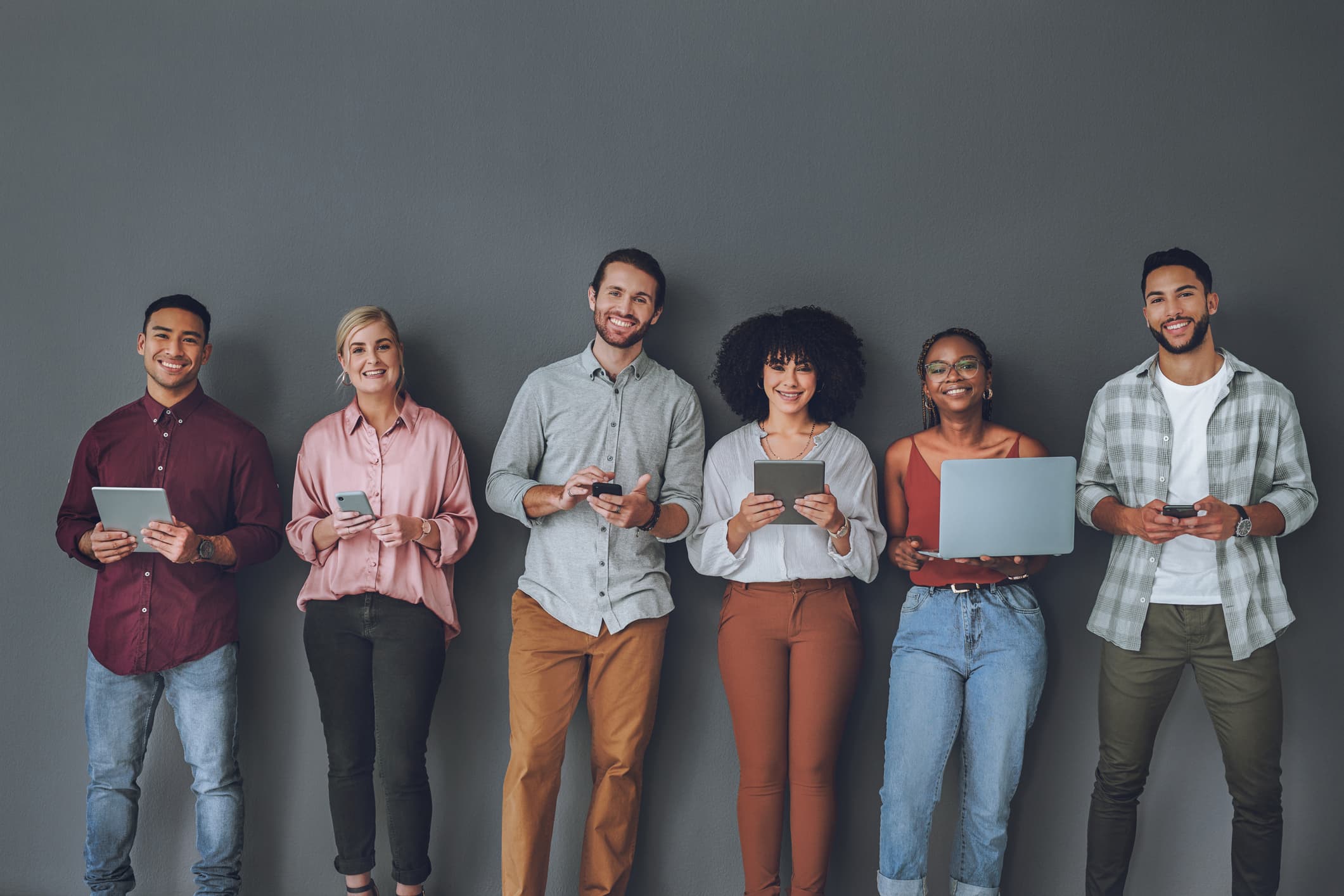 Six people stood up holding digital devices and smiling