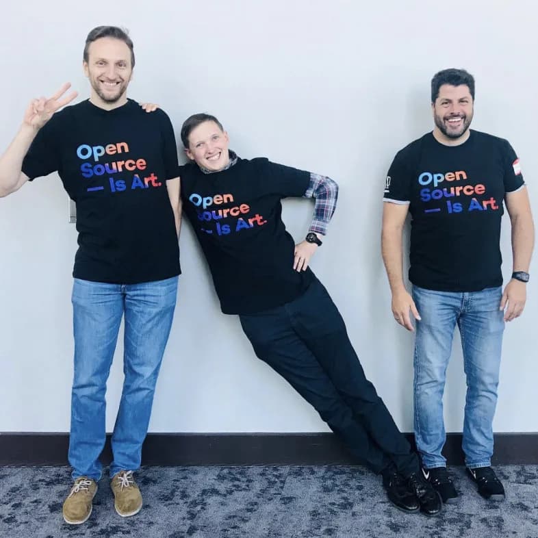 Three people in matching 'Open Source — Is Art.' shirts, smiling, with one playfully leaning.