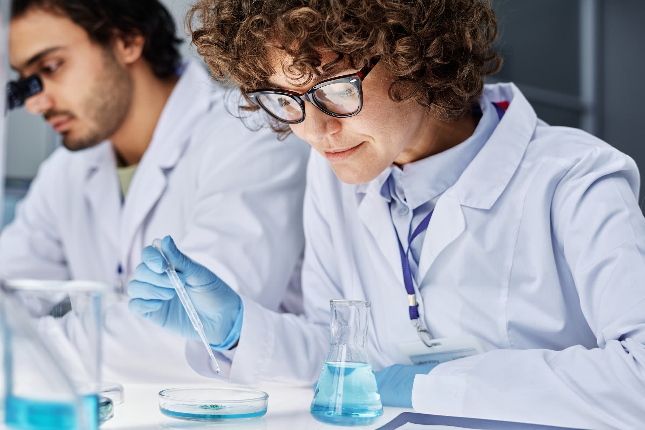 A bespectacled female pharmacist holds a pipette in her right hand and squeezes liquid into a petri dish, while a male pharmacist behind her looks into a microscope.